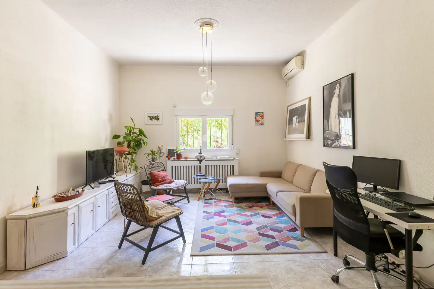 Bright living room with beige sectional, colorful rug, desk, and rattan chairs. White walls and tile floor.