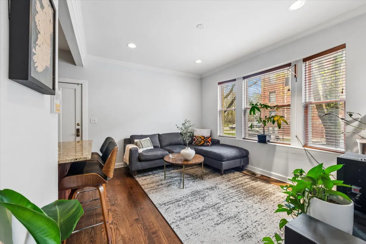 Living room with a gray sectional sofa, round wood table, and patterned rug. Three windows with blinds let in natural light. Plants add a touch of greenery.