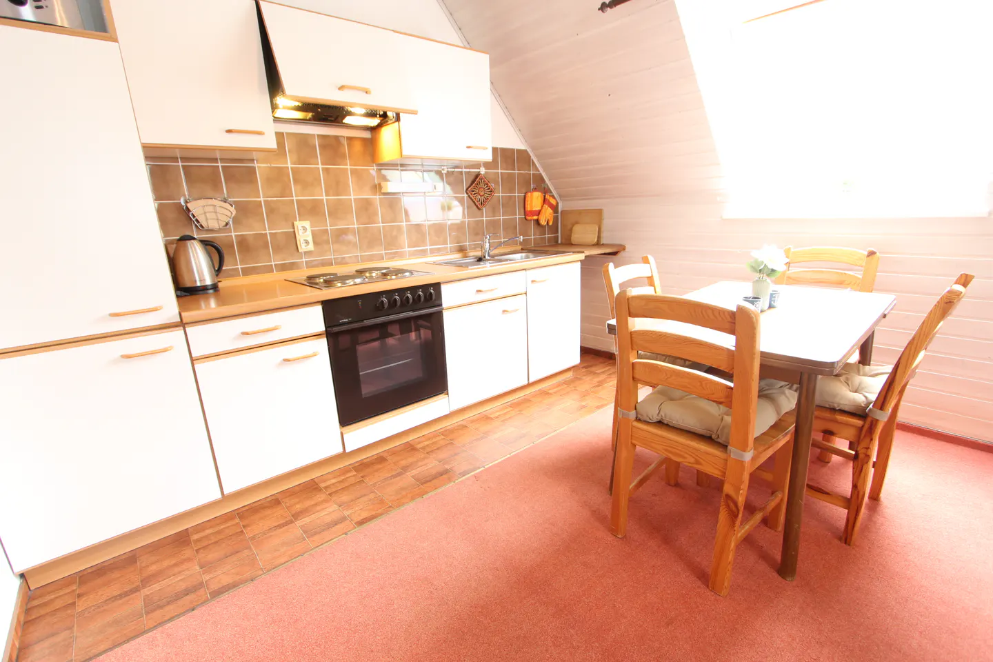 Bright kitchen with white cabinets, brown tile backsplash, and a black oven. A wooden table and chairs sit on a pink carpet.