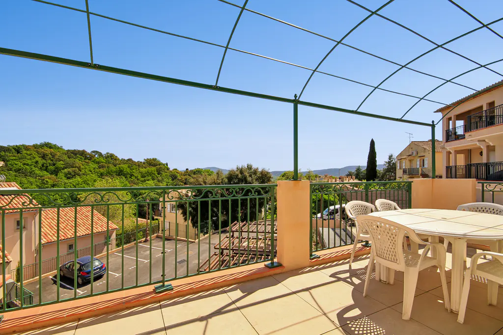 Outdoor patio with green metal railing, beige tile floor, and white plastic table and chairs. View of trees, parking lot, and blue sky.