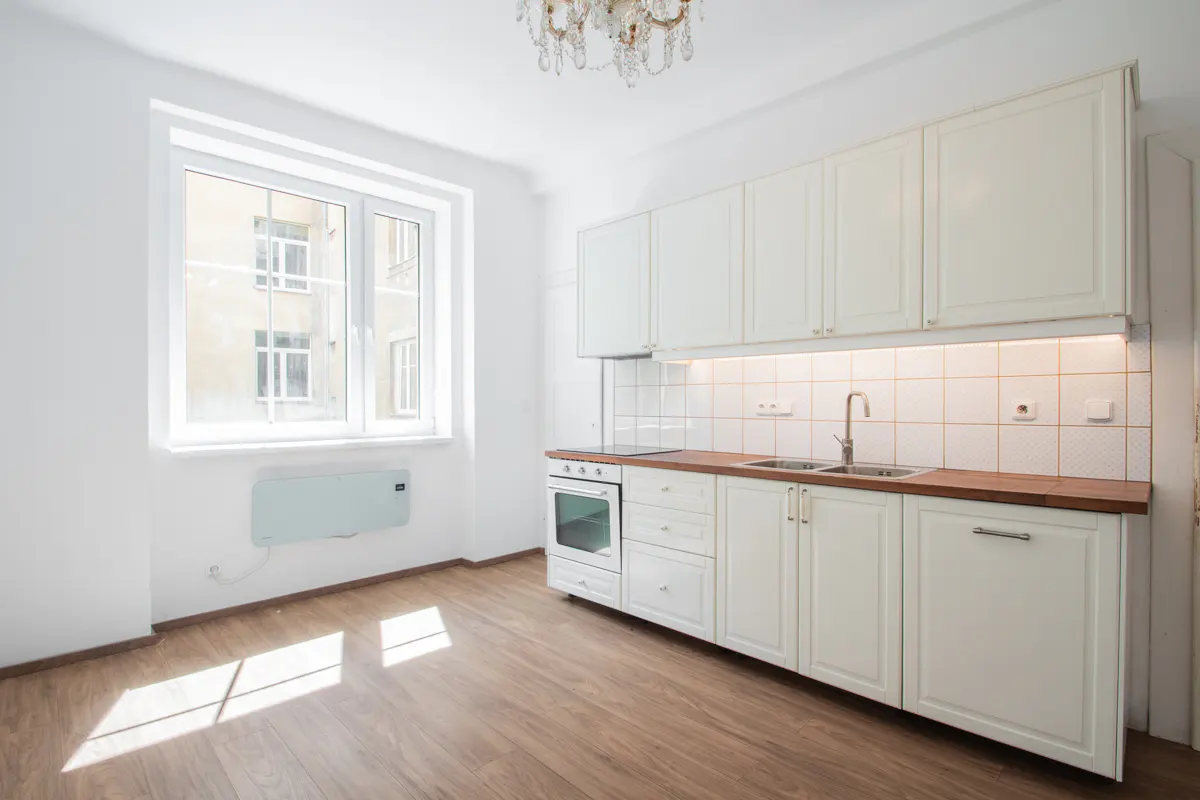 Bright, empty kitchen with white cabinets, wood countertops, and a chandelier. Sunlight streams through a window onto the wood floor.