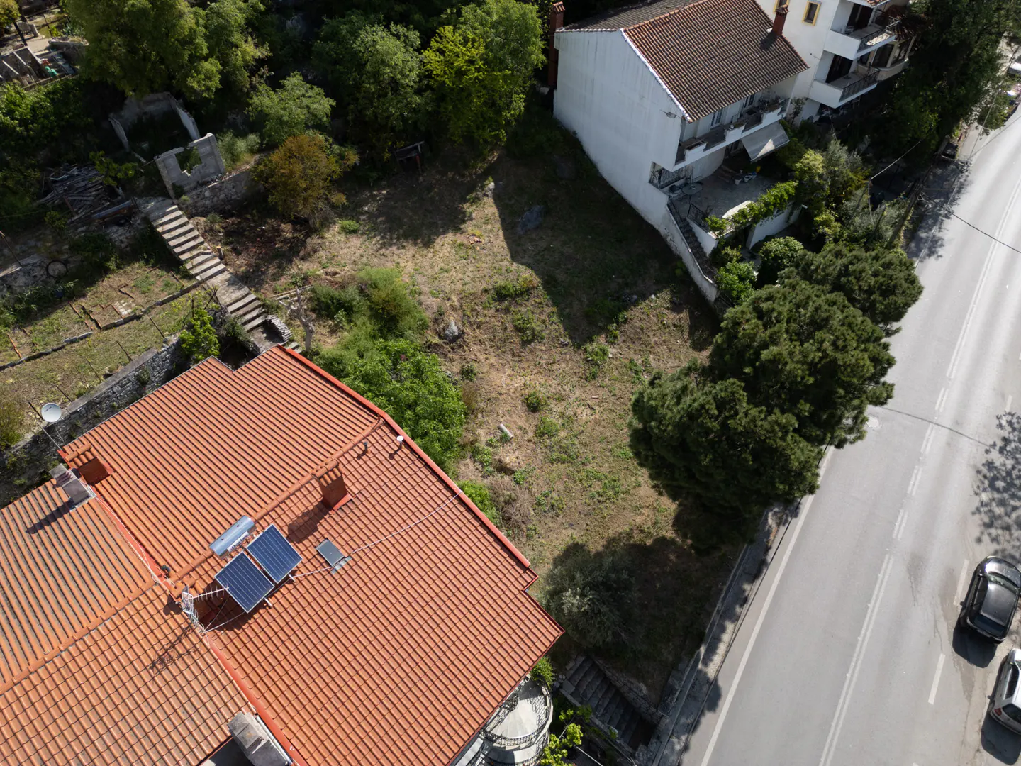 Aerial view of a vacant lot between houses, with a road on the right and trees in the background. A house with solar panels is in the foreground.