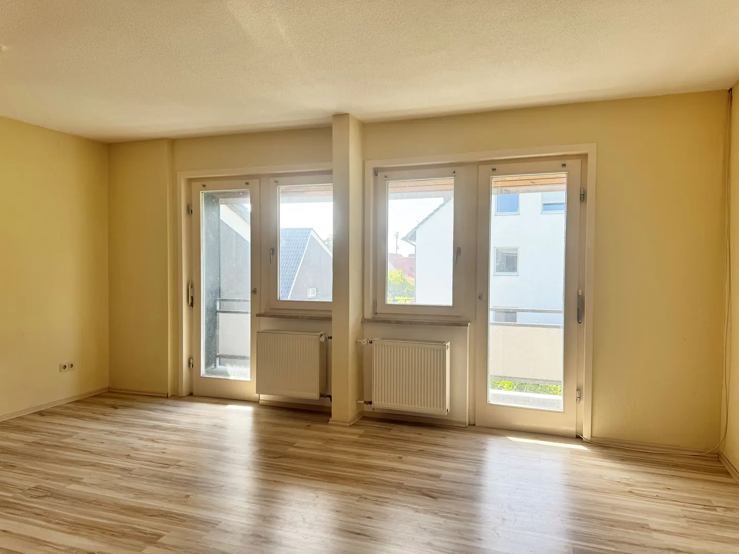 Bright, empty room with wood-look floors, cream walls, and two sets of white-framed glass doors leading to balconies. Radiators are under each window.
