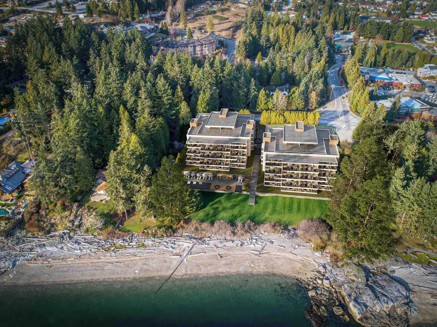 Aerial view of two modern, multi-story condos near a beach and forest. The buildings have balconies and a green lawn. The water is turquoise.