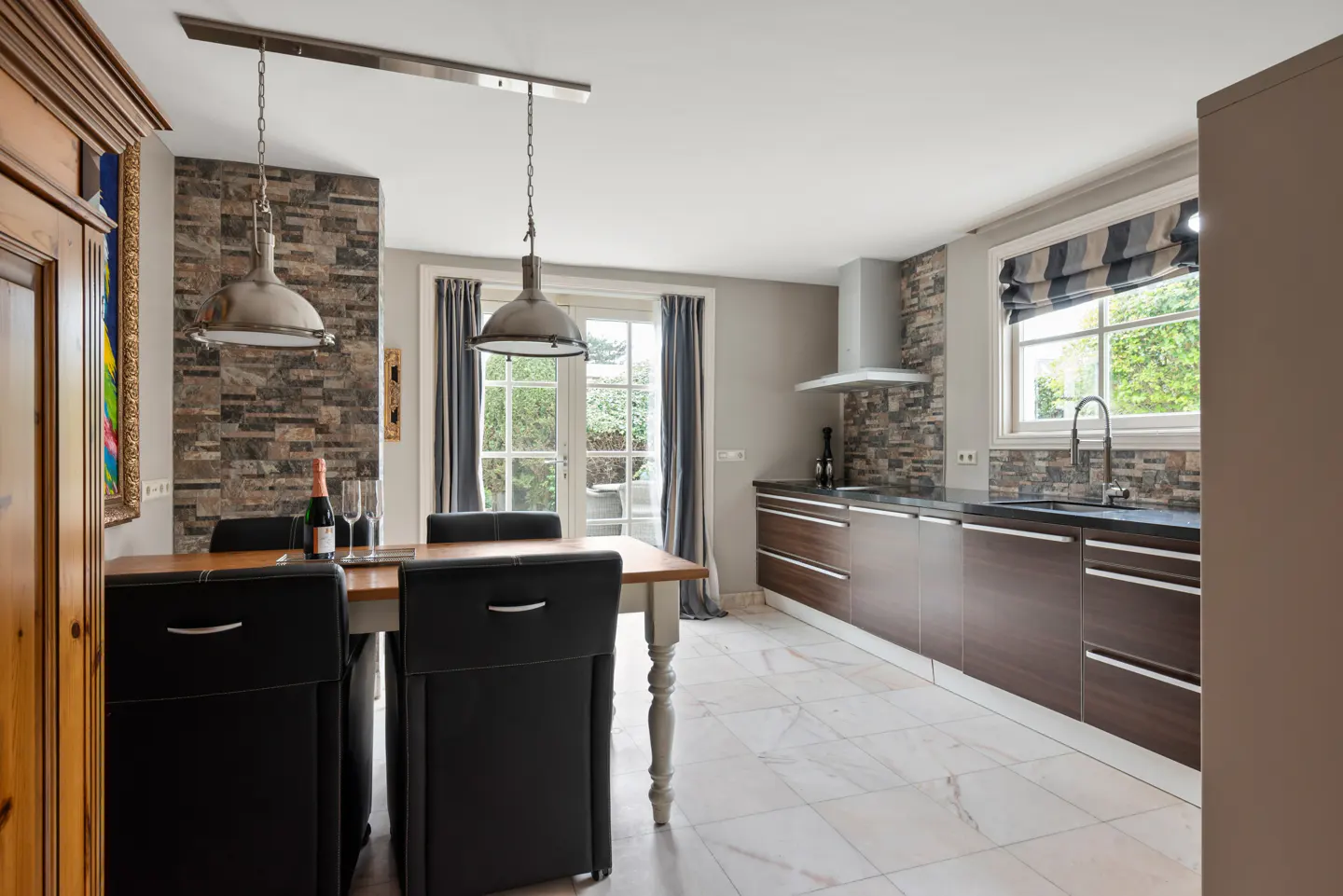 A modern kitchen with white marble floors, brown cabinets, and a wooden table with black chairs. Two pendant lights hang above the table. A bottle of wine and glasses sit on the table.