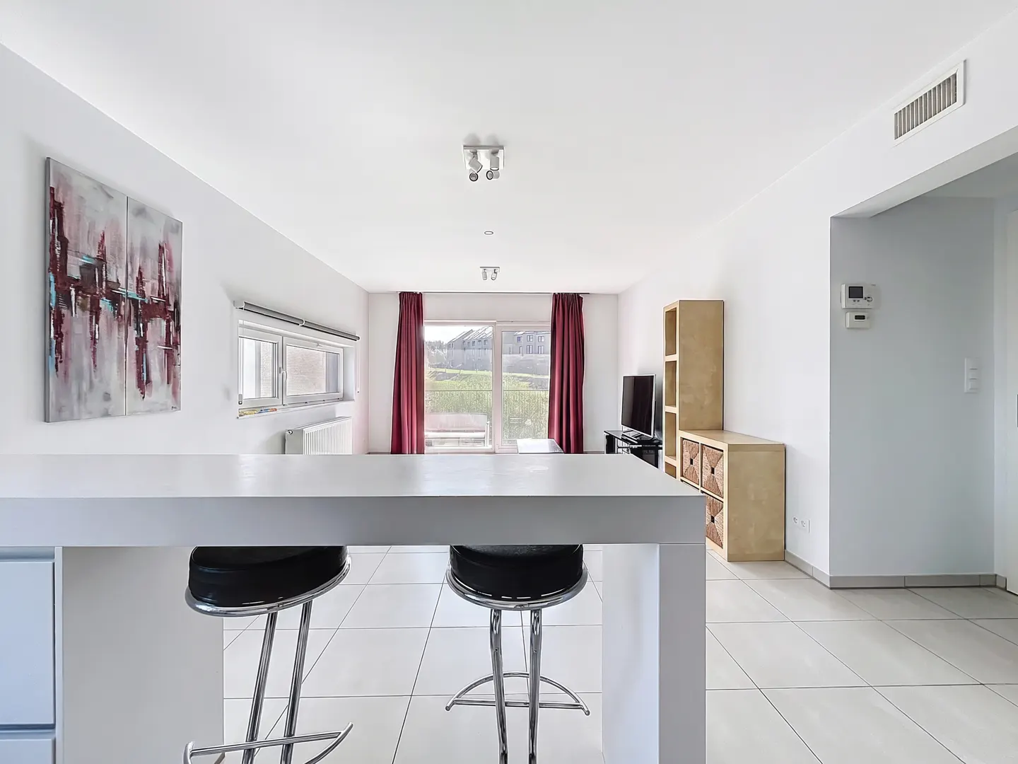 Bright, modern living room with white walls, tile floors, and a white breakfast bar with two black stools. Abstract art hangs on the wall.
