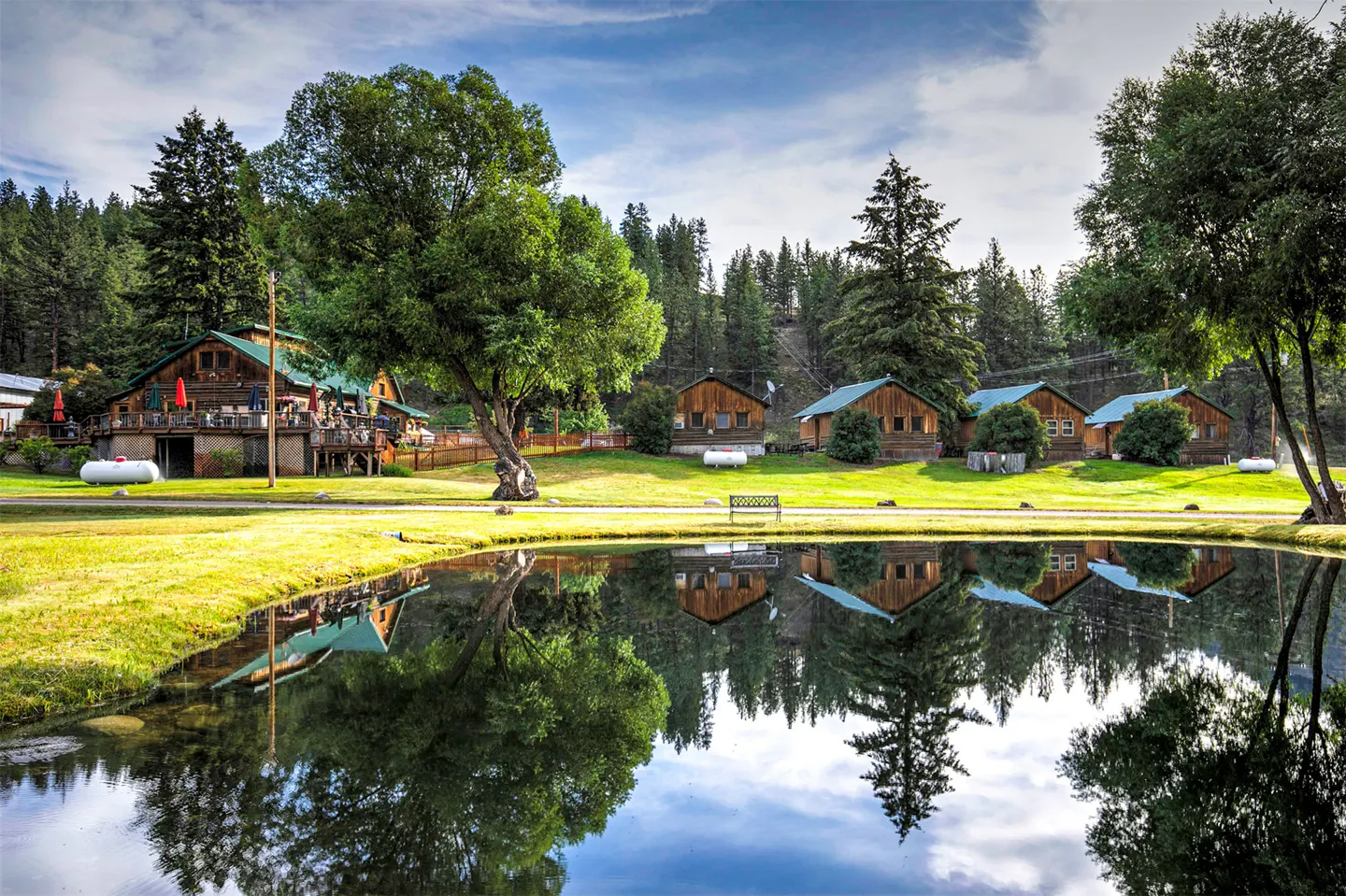 Scenic view of rustic cabins reflected in a pond, surrounded by green grass and trees under a blue sky.
