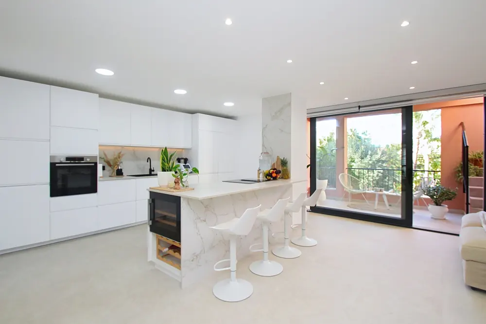 Bright, modern kitchen with white cabinets, marble island with four white stools, and sliding glass doors to a balcony.