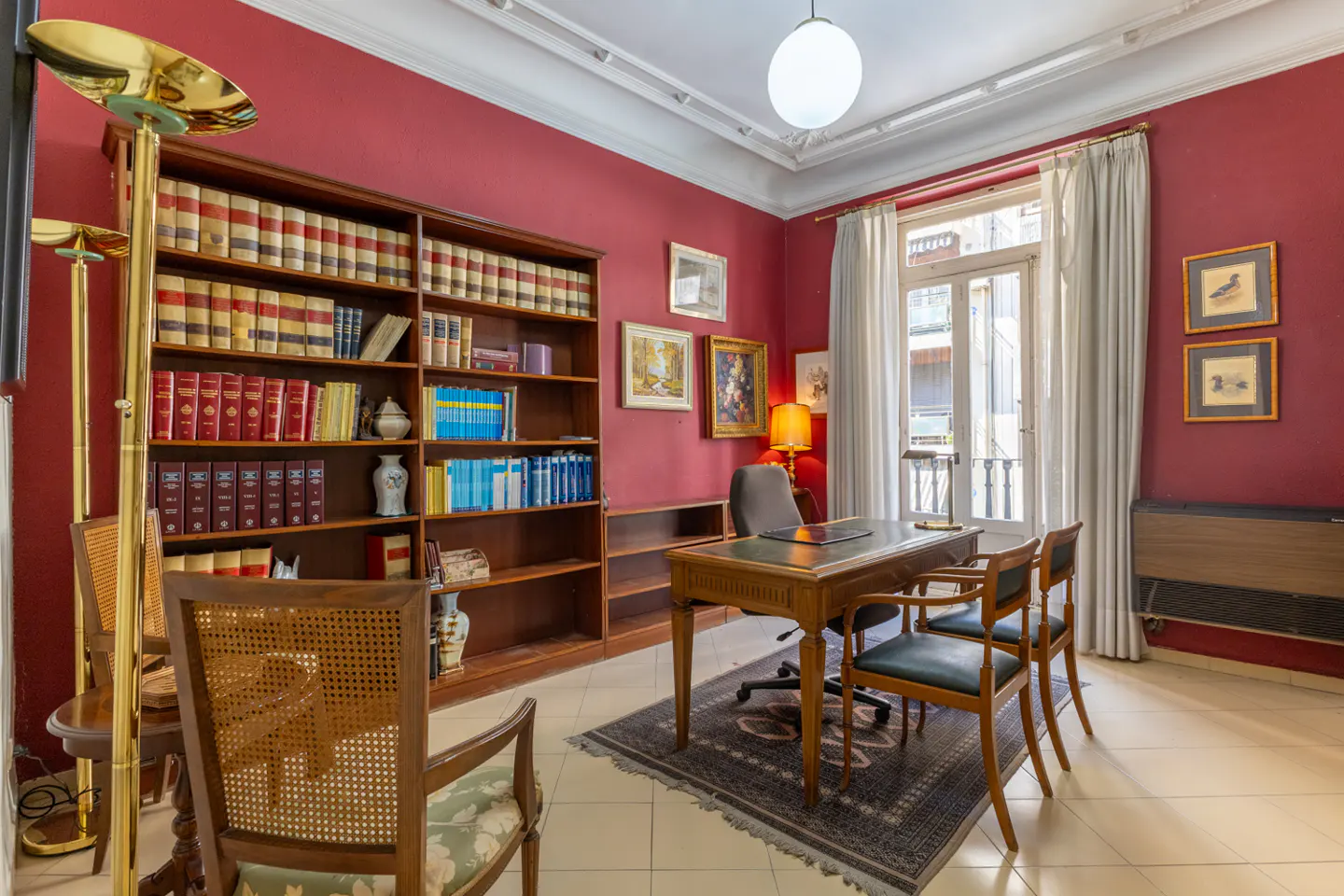 A home office with red walls, a large bookcase filled with books, and a wooden desk with chairs.
