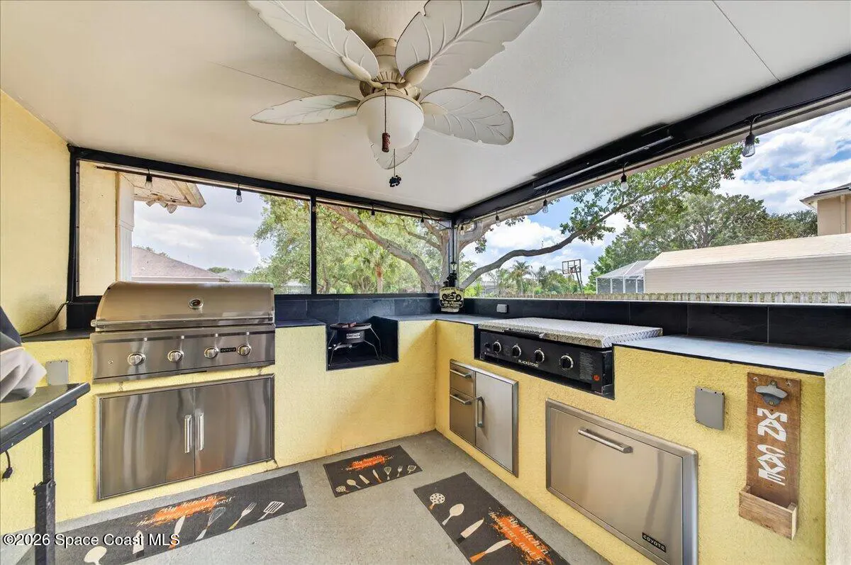 Outdoor kitchen with stainless steel grill, black countertops, and yellow stucco walls. A leaf-shaped ceiling fan hangs above.