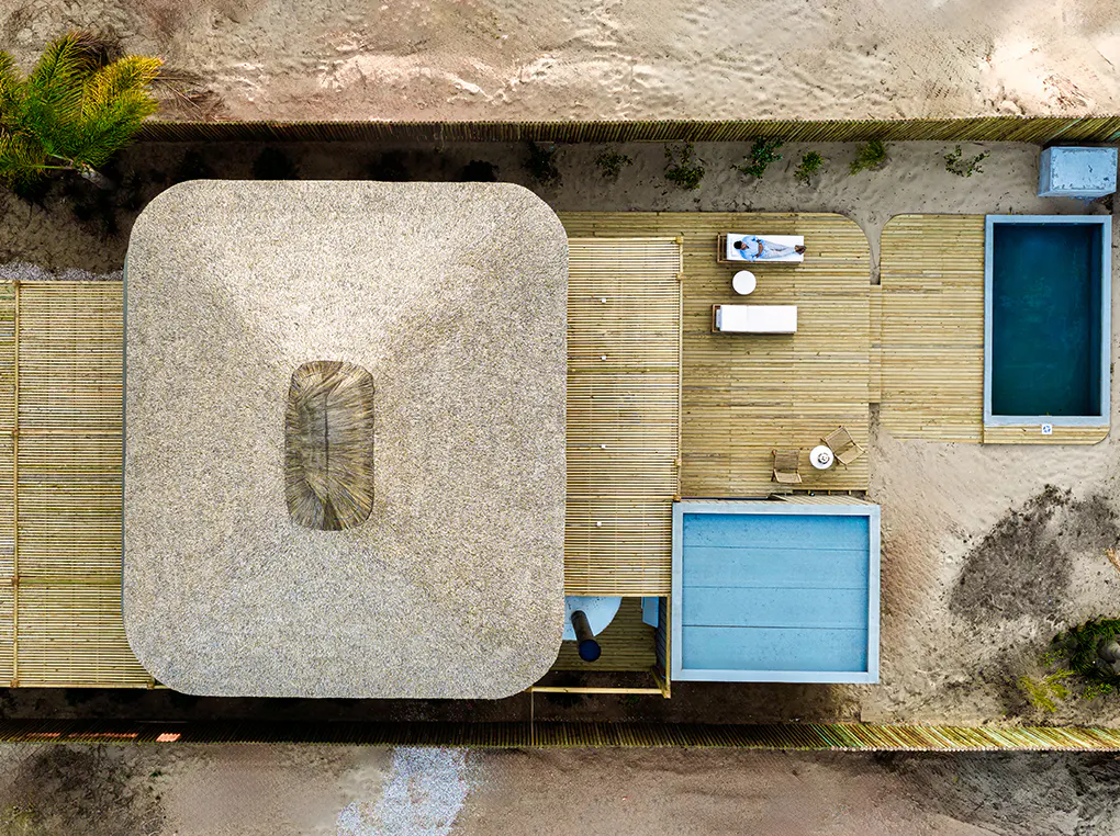 Aerial view of a thatched-roof house with a wooden deck, two pools, and a person relaxing on a lounge chair.