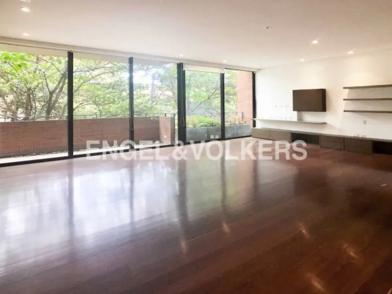 An empty living room with dark wood floors, white walls, and large windows overlooking greenery. A TV and shelves are mounted on the wall.