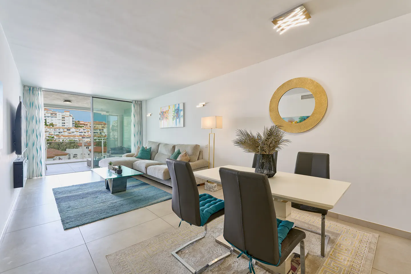 Bright, modern living room with a beige sectional sofa, glass coffee table, and dining area with a white table and gray chairs. Balcony view of a coastal town.