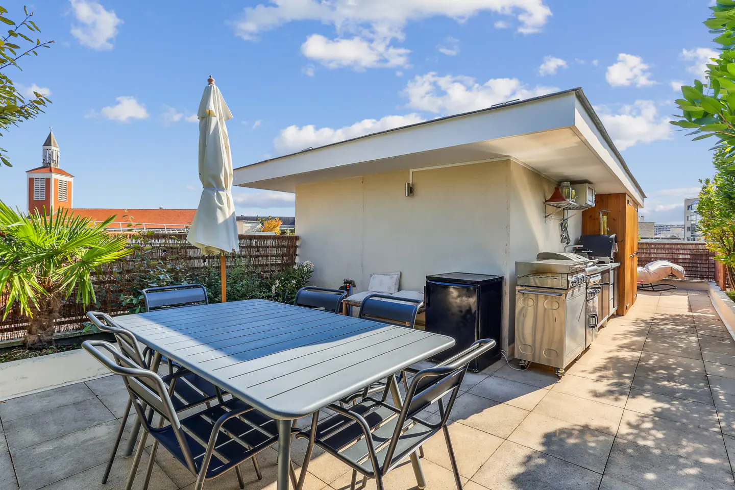 Rooftop patio with a gray table, six chairs, a grill, and a mini-fridge under a blue sky with white clouds.