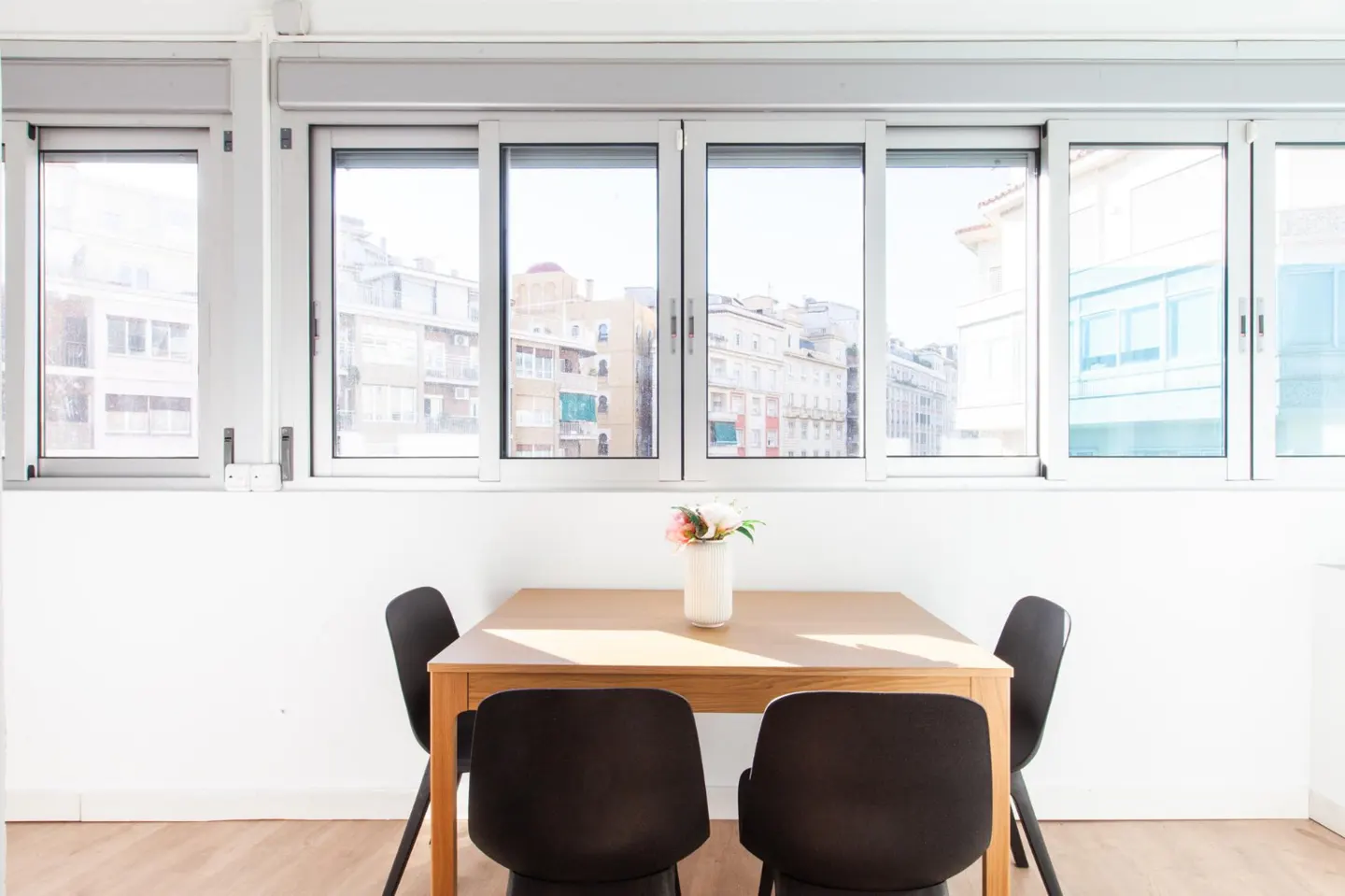 Bright dining area with a wooden table, four black chairs, and a vase of flowers. Large windows offer a city view.