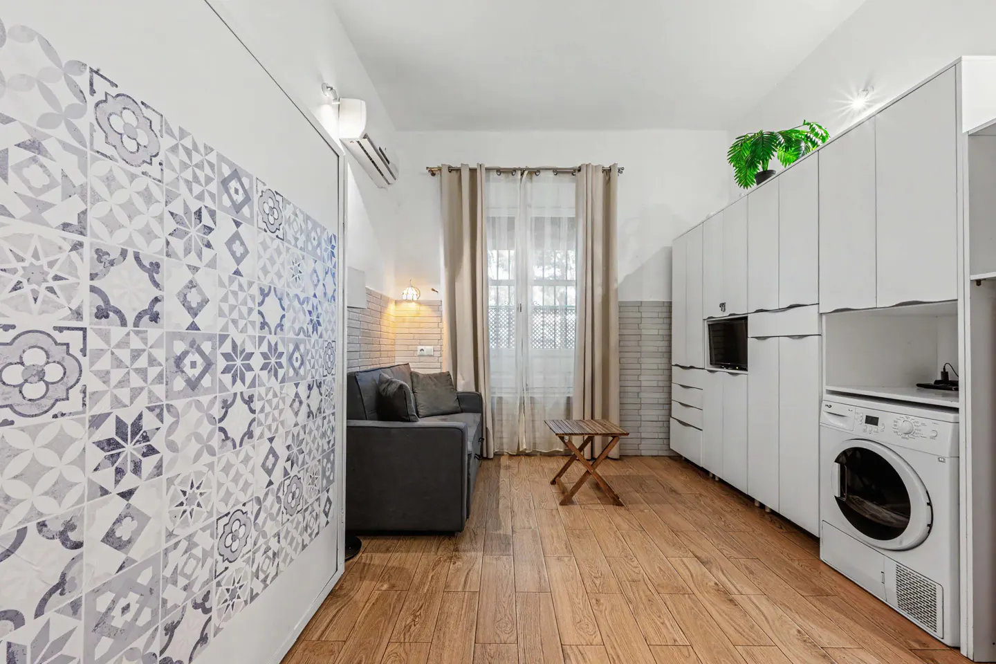 Cozy apartment interior with wood floors, a gray sofa, and white cabinets with a washing machine. A blue-patterned wall adds a unique touch.