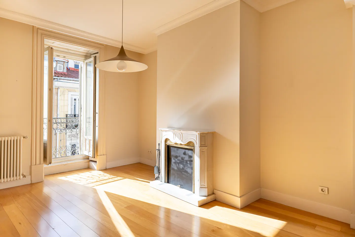 Bright, empty room with wood floors, cream walls, and a white marble fireplace. Open balcony doors show a European city view.