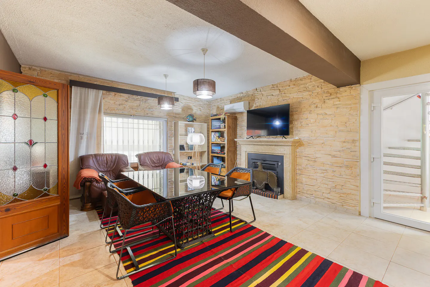 Living room with stone wall, fireplace, TV, glass table, wicker chairs, and colorful striped rug. Brown leather chairs are near a window.