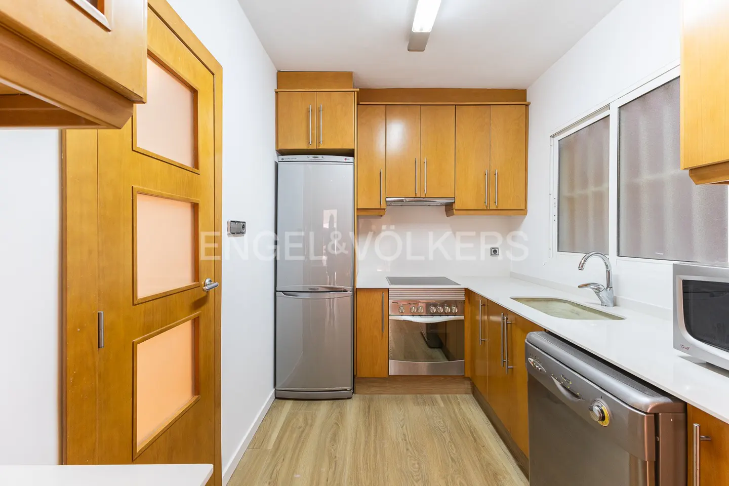 Bright kitchen with wood cabinets, stainless steel appliances, and a wood door with frosted glass panels. White countertops and walls.