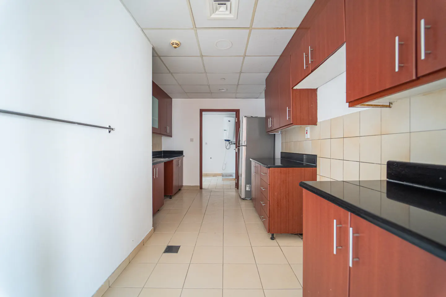 A kitchen with brown cabinets, black countertops, and beige tile flooring. A stainless steel refrigerator is visible.