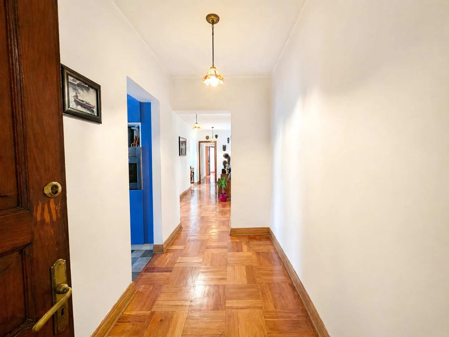 A bright hallway with parquet floors leads to other rooms. A wooden door is ajar on the left. White walls and a pendant light fixture.