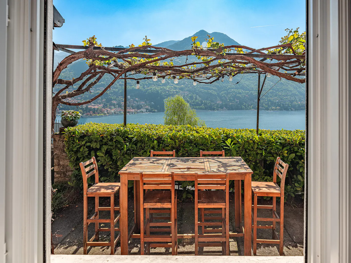 Outdoor patio with wooden table and chairs, viewed through a doorway. A vine-covered trellis hangs above, with a lake and mountains in the background.