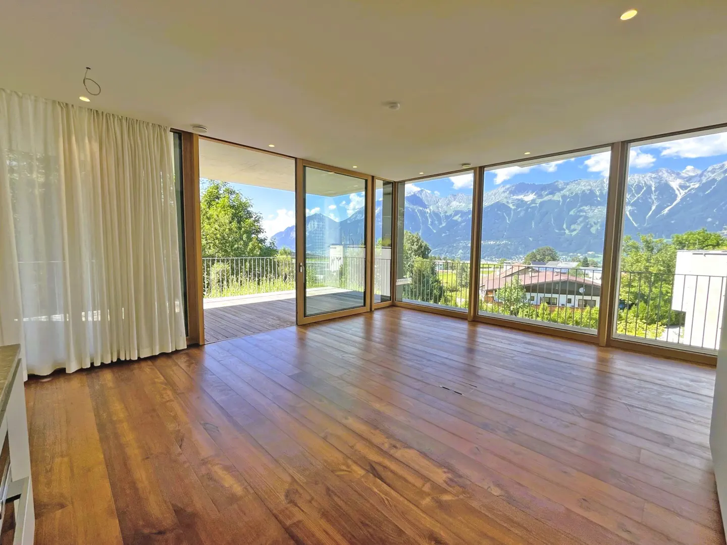 Bright, empty room with wood floors and large windows showing mountain views. White curtains hang to the left.