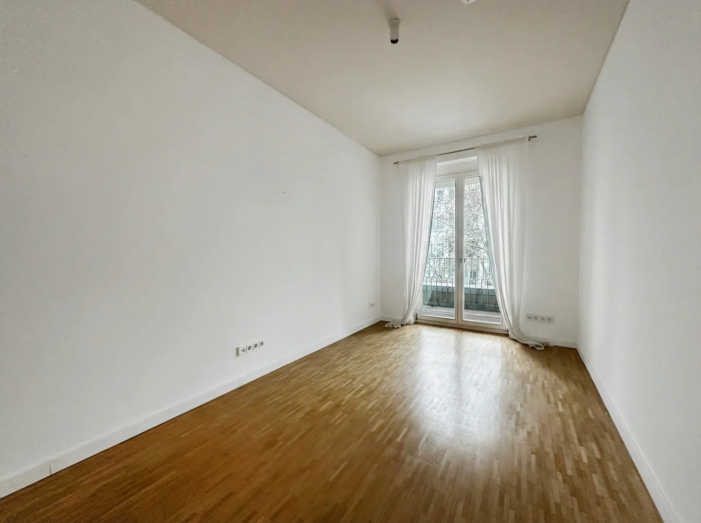 Empty room with white walls, light wood flooring, and a glass door leading to a balcony with white curtains.