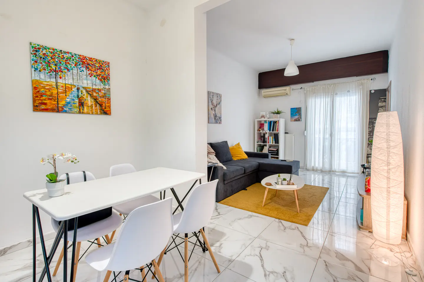 Bright, modern apartment interior with white walls, marble floors, and open-plan living. Dining table with white chairs, colorful artwork, and a gray sofa with yellow accents.