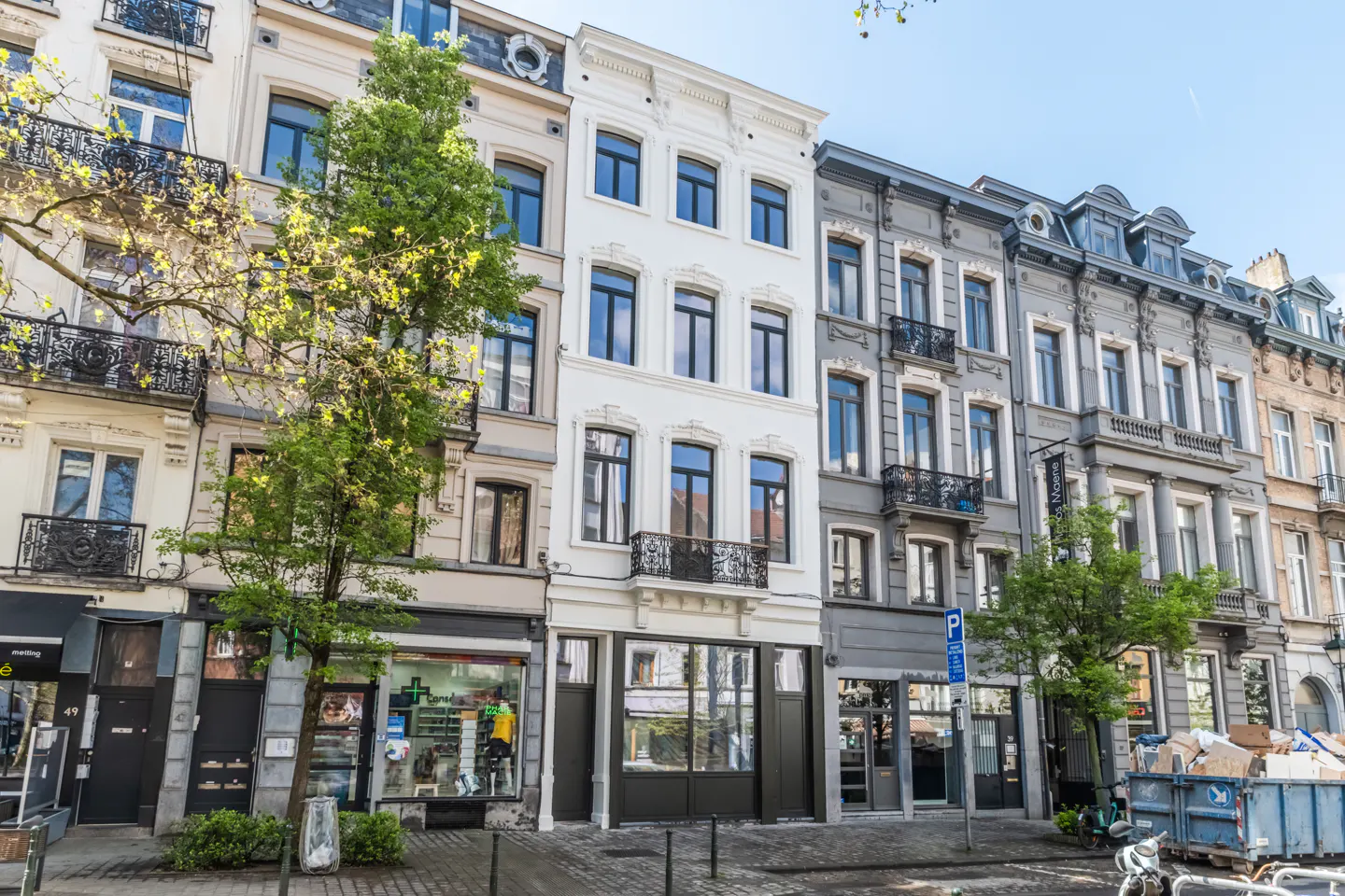 Row of multi-story buildings with shops on the ground floor, varying in color from beige to gray, with trees.