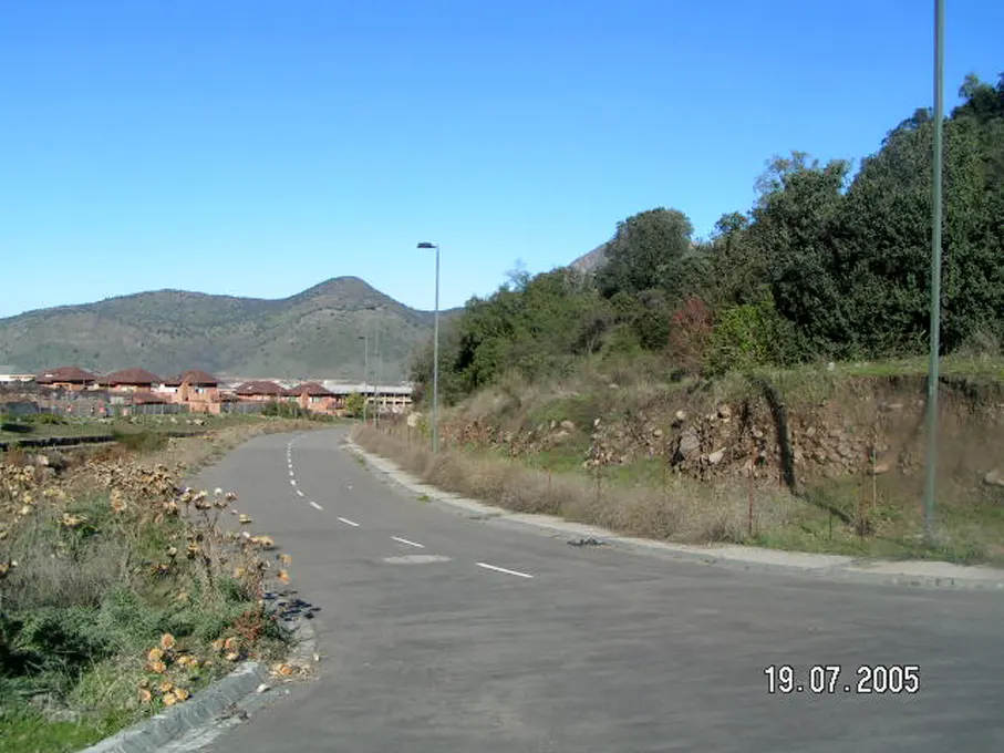 Curving asphalt road with white lines, bordered by dry vegetation and a hillside with trees. Houses and a mountain are in the background under a blue sky.