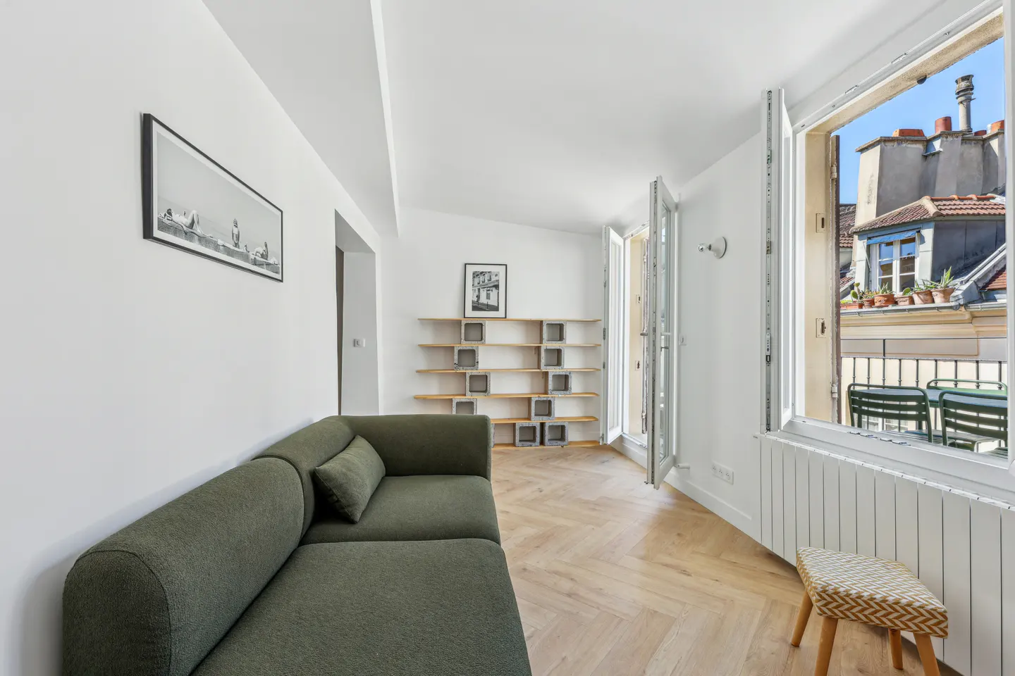 Bright living room with a green sofa, wooden floors, and a balcony view of rooftops.