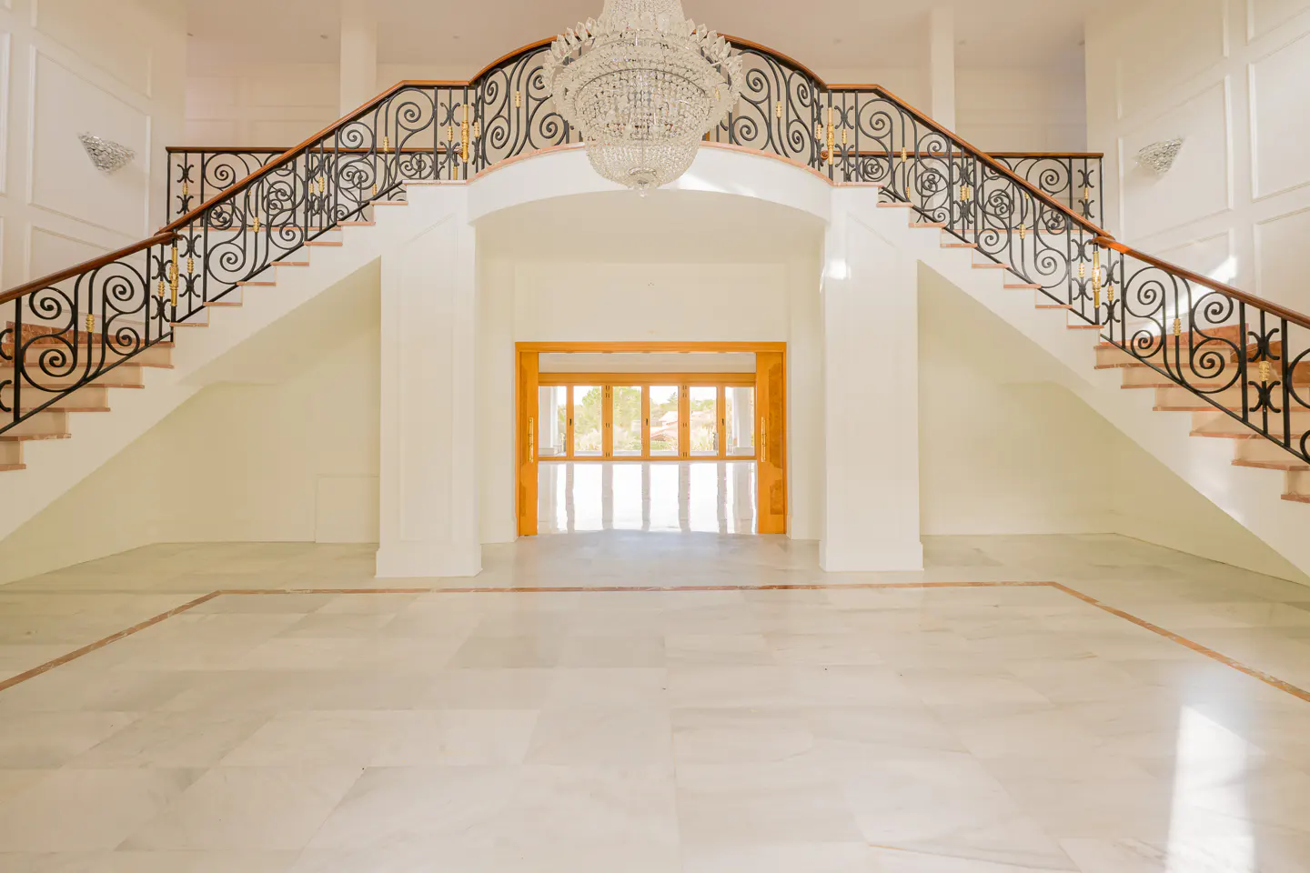Bright, white foyer with marble floors, split staircase with black wrought iron railings, and a crystal chandelier.
