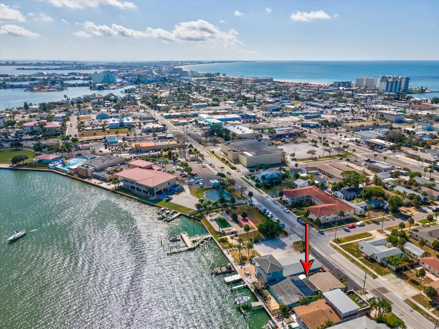 Aerial view of a waterfront property with a blue house and a red arrow pointing to it, near a road and a body of water.