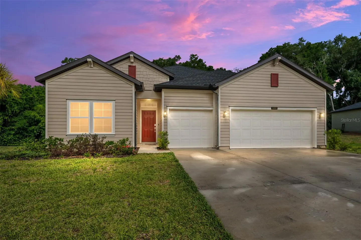Tan siding house with a red door, two-car garage, and green lawn under a pink and purple sky.