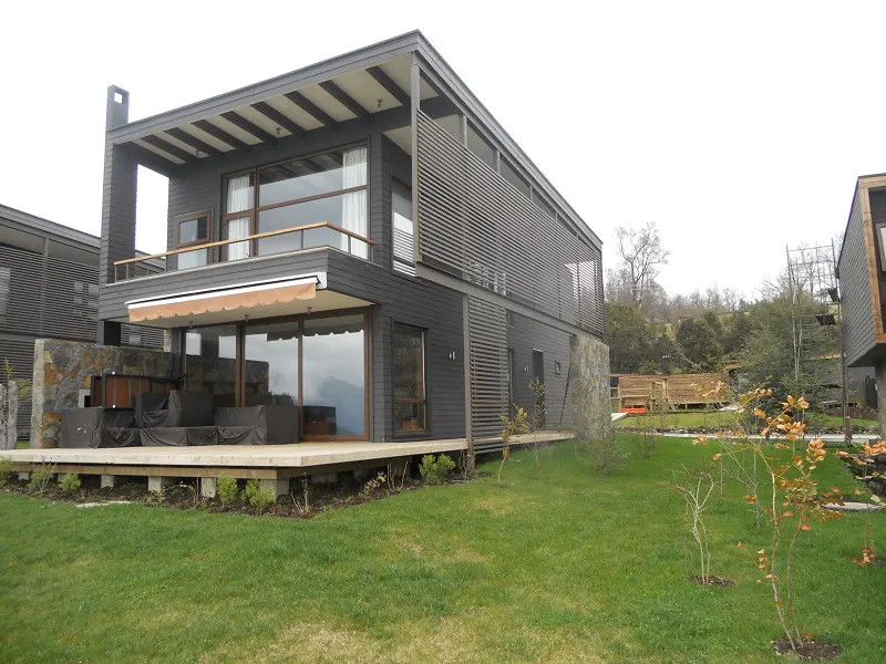 Modern two-story house with gray siding, large windows, and a wooden deck with covered furniture on a green lawn.