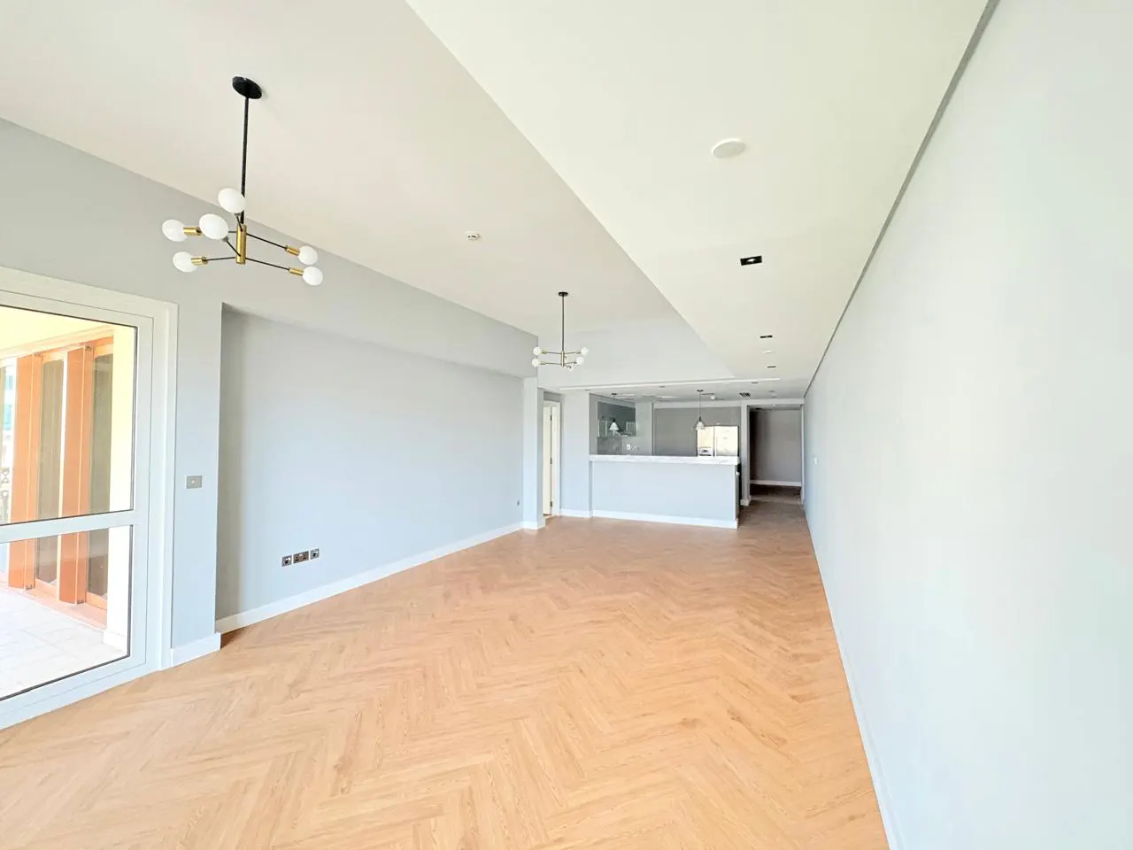 Bright, empty living room with herringbone wood floors and light gray walls. Modern chandeliers hang from the white ceiling. A doorway leads to a kitchen area.