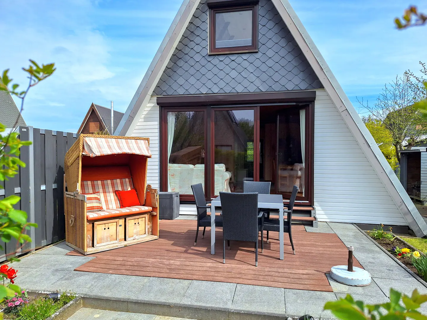 A-frame house with a patio, a wicker beach chair with red cushions, and a table with four chairs.
