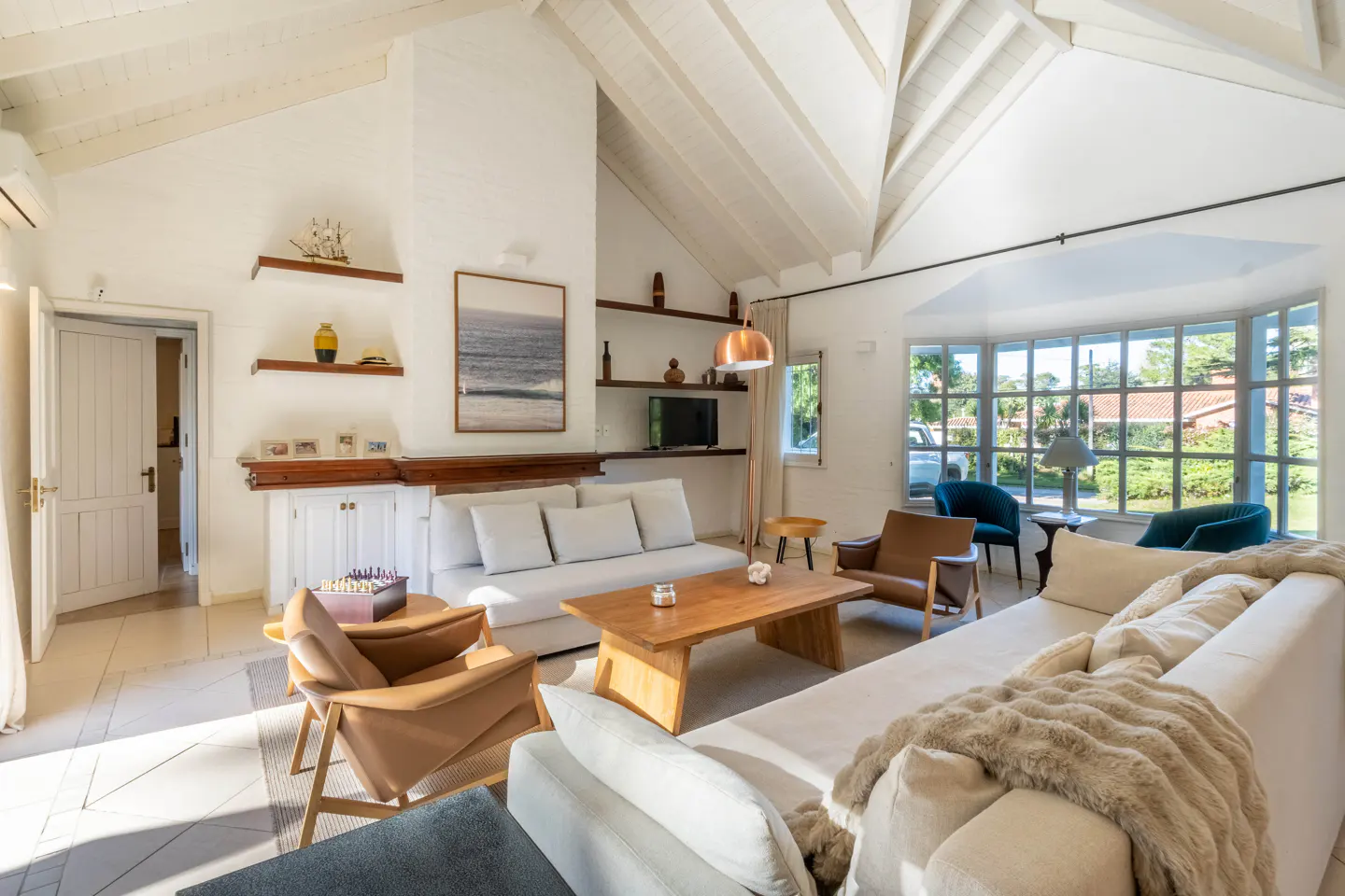 Bright living room with white walls, vaulted ceiling, and wood beams. Sofas, chairs, wood table, shelves, and a large window with a view.