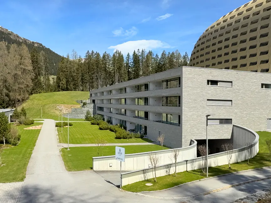Modern grey apartment building with balconies, green lawn, and parking sign. Forest and blue sky in the background.