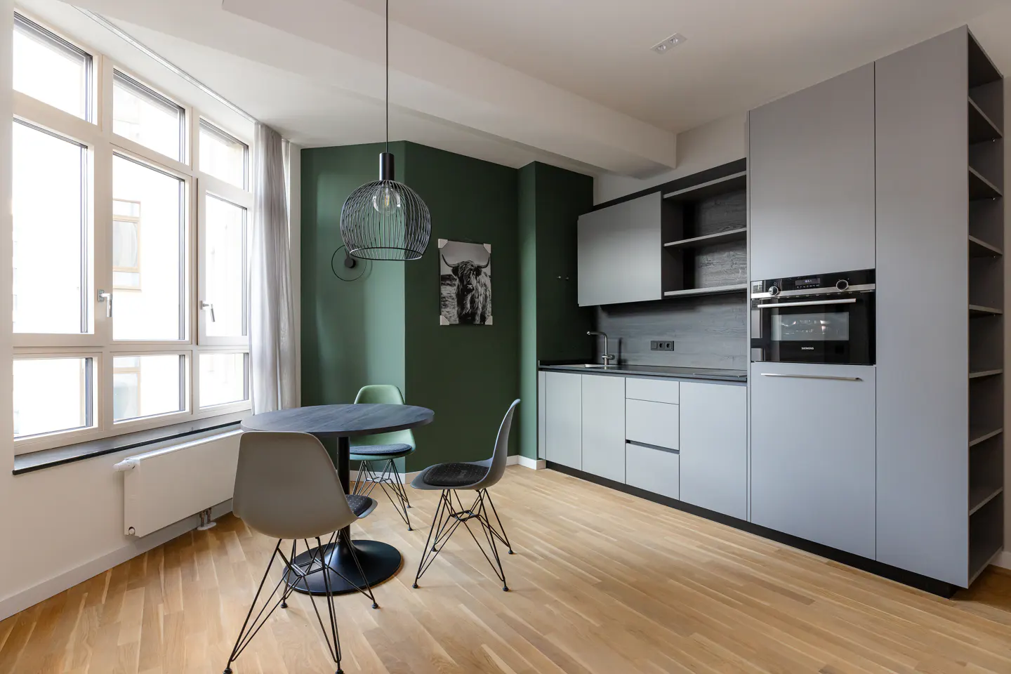 A modern kitchen with gray cabinets, a round table with chairs, and a large window. The walls are painted a dark green.