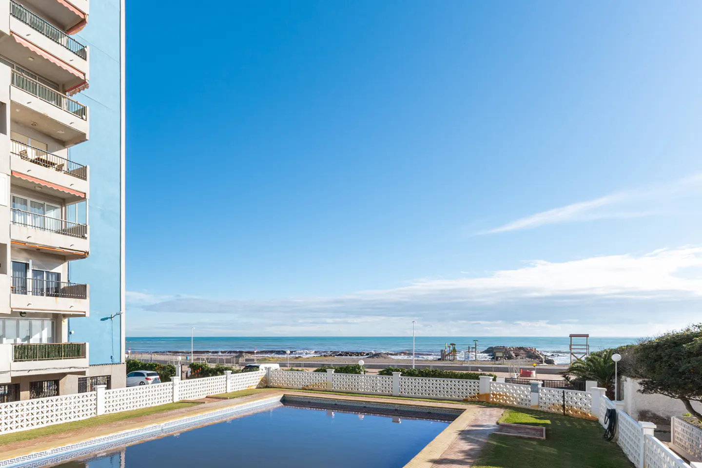 View of a swimming pool in the foreground with a high-rise apartment building and the ocean in the background.