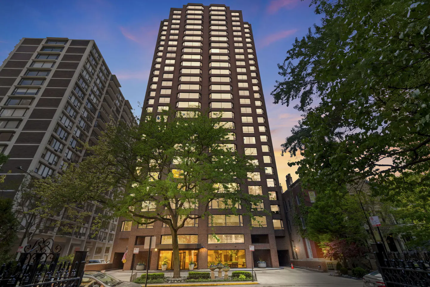 Exterior view of a tall, brown brick apartment building with many windows, trees, and a blue and orange sky.