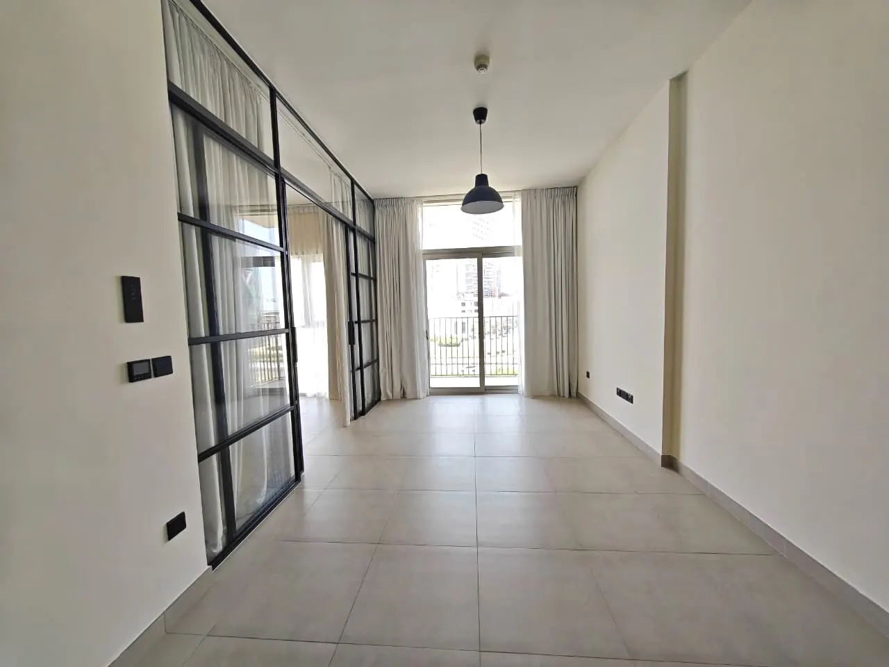 Empty, bright room with white walls, gray tile floor, and black-framed glass doors leading to a balcony with white curtains. A black pendant light hangs from the ceiling.