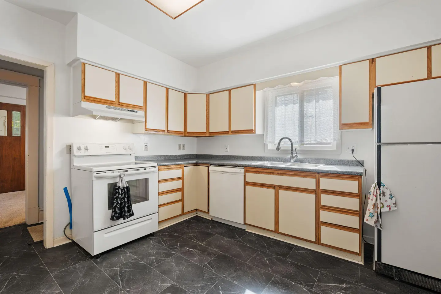 Kitchen with white appliances, light cabinets with wood trim, dark marble-look tile floors, and a window over the sink.