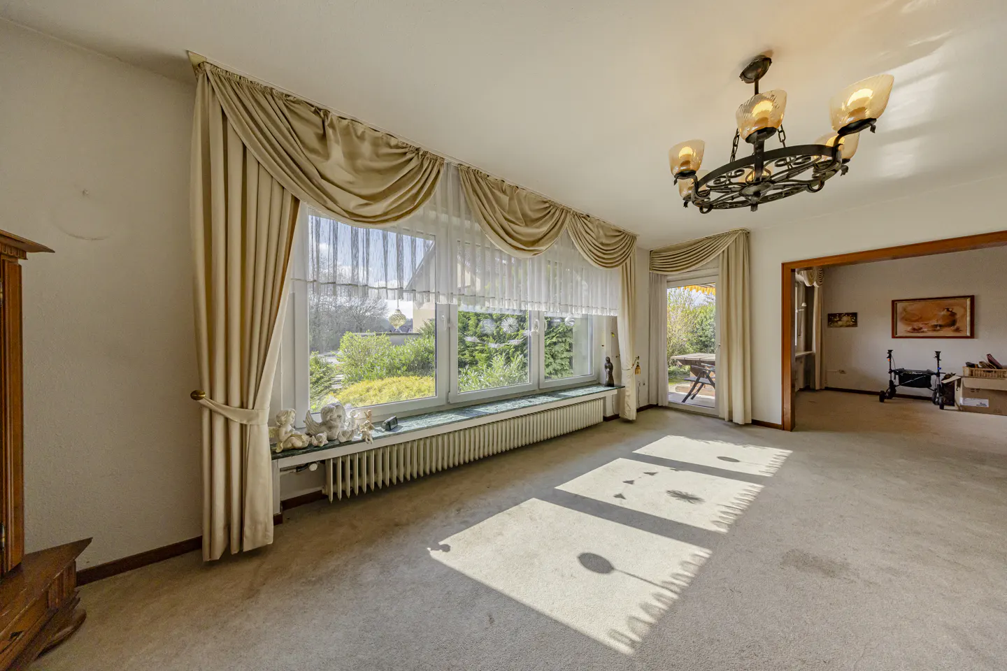 Bright living room with beige carpet, large window with curtains, and a wrought iron chandelier. Sunlight streams across the floor.