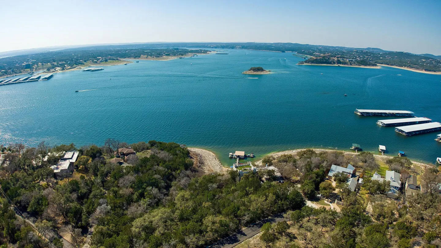 Aerial view of a blue lake with green trees, houses, and docks along the shoreline under a clear blue sky.