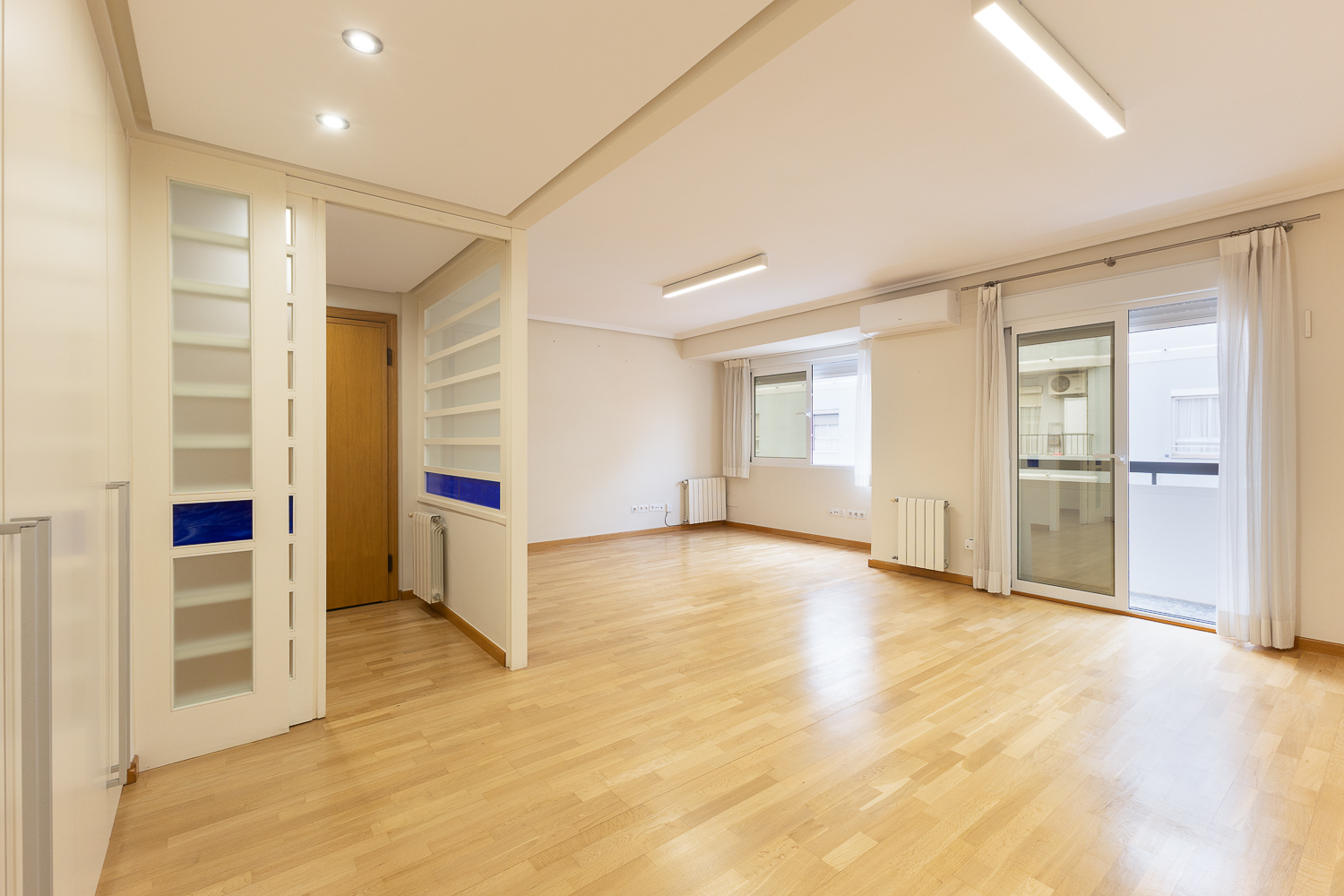 Bright, empty living room with light wood floors, white walls, large windows, and a glass-paneled room divider.
