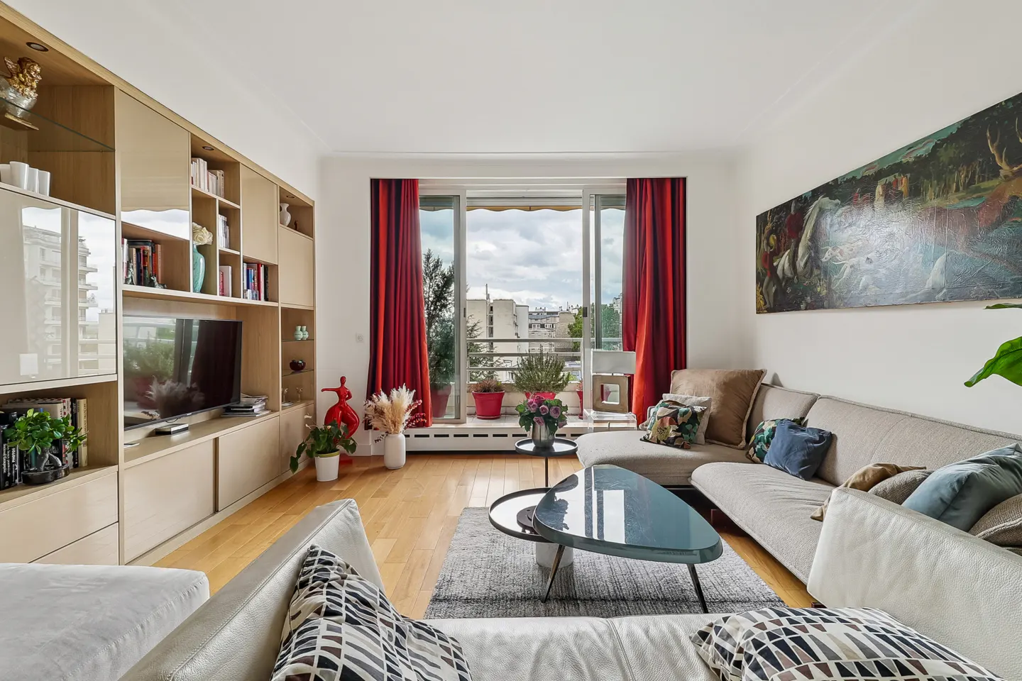 Bright living room with hardwood floors, built-in shelving, and a large window with red curtains. A grey sofa and glass coffee table sit on a patterned rug.