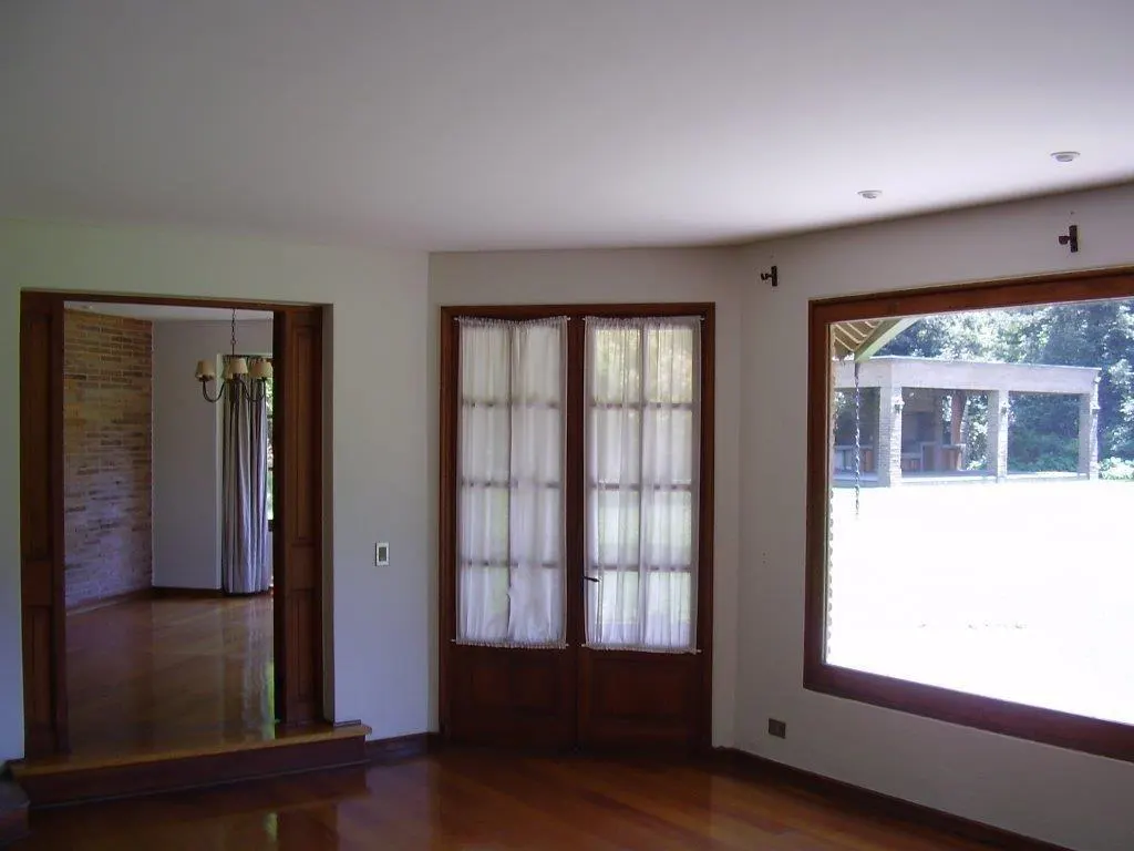 A bright room with hardwood floors, white walls, and wood trim. A doorway leads to another room with a brick wall. A window and glass doors look out to a yard.